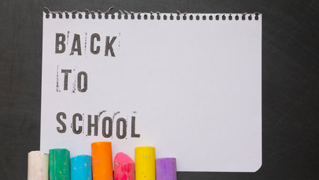 blank page of a notebook with the phrase "Back To School" written in English with worn up letters next to colored chalk on a black chalkboard backgroundの写真素材