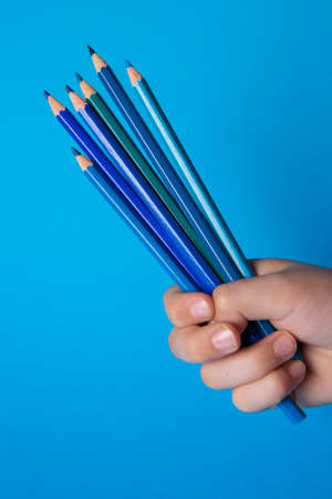 Hand of a child holding wooden pencils of different shades of blue color on blue backgroundの写真素材