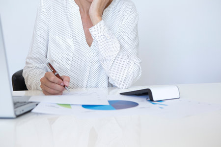 Middle-aged woman in white shirt analyzing statistics and graphs with her computerの写真素材