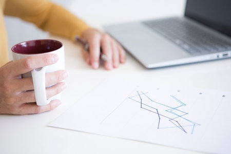 Hands of a woman in mustard yellow shirt drinking a cup of tea while working with her mobile and computer in financeの写真素材