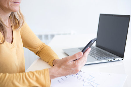 Hands of a woman in mustard yellow shirt holding her mobile phone while analyzing financial statistics and graphsの写真素材