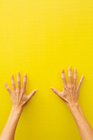 Vertical photograph of both hands and arms of a woman showing all the fingers of each hand with the nails painted yellow on a yellow background.の写真素材