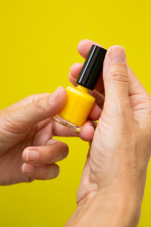 Vertical photograph of a woman's hand with yellow nails holding a bottle of yellow nail polish on a yellow background.の写真素材
