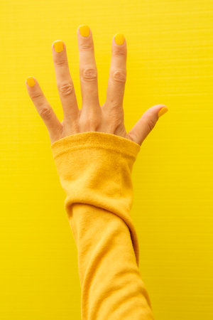 Vertical photograph of a woman's hand with her nails painted yellow on a yellow background showing all five fingers.の写真素材