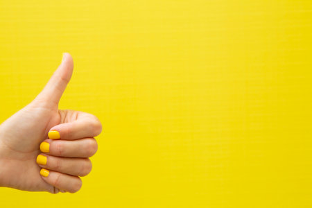 Horizontal photograph of a woman's hand with her nails painted yellow, giving a thumbs-up gesture on a yellow background.の写真素材