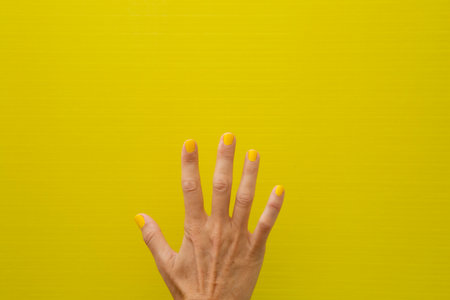 Horizontal photograph of a woman's hand with her nails painted yellow on a yellow background showing all five fingersの写真素材