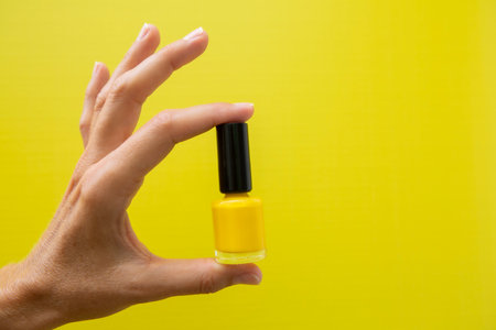 Horizontal photograph of a woman's hand with yellow nails holding a bottle of yellow nail polish on...