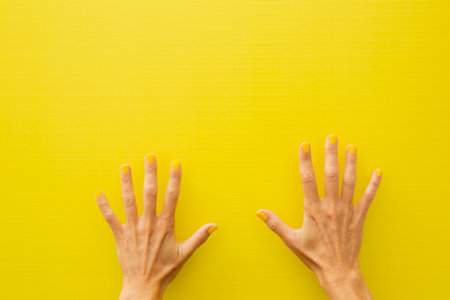 Horizontal photograph of a woman's two hands showing all the fingers of each hand with the nails painted yellow on a yellow backgroundの写真素材