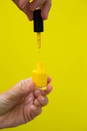 Vertical photograph of the drop of yellow nail polish falling on the bottle while being held by a woman's hand on a yellow background.の写真素材