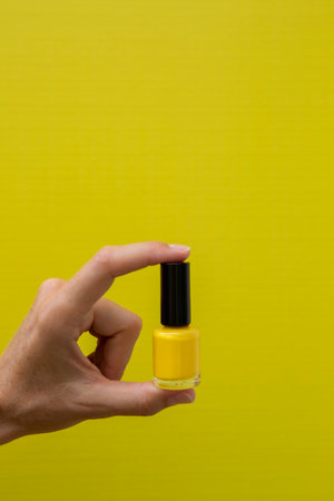 Vertical photograph of a hand with yellow nails holding a bottle of yellow nail polish on a yellow background.の写真素材