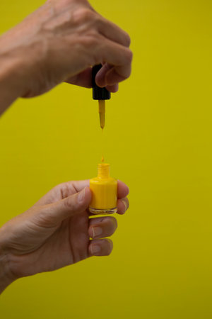 Vertical photograph of the drop of yellow nail polish falling on the bottle while being held by a woman's hand on a yellow background.の写真素材