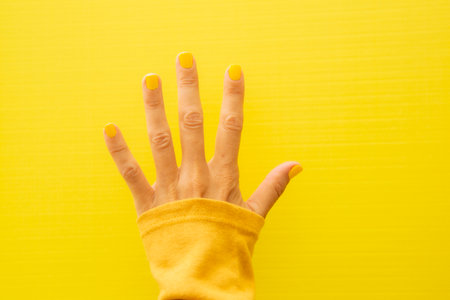 Horizontal photograph of a woman's hand with her nails painted yellow on a yellow background showing all five fingers.の写真素材