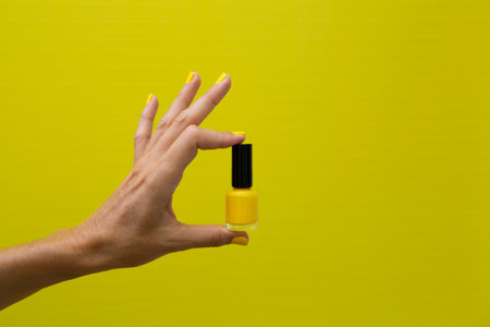 Horizontal photograph of a woman's hand with yellow nails holding a bottle of yellow nail polish on a yellow background.の写真素材