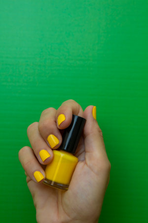 Vertical photograph of a woman's hand with unpainted nails holding a bottle of yellow nail polish in front of a green background.の写真素材