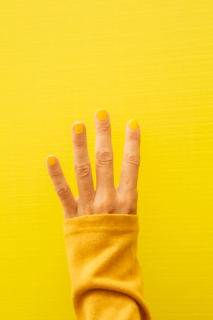 Vertical photograph of a woman's hand with her nails painted yellow on a yellow background showing all four fingers.の写真素材