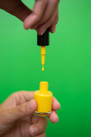 Detail of the drop of yellow nail polish falling on the bottle while being held by a woman's hand against a green background.の写真素材