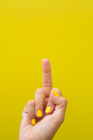 Vertical photograph of a woman's hand with her nails painted yellow, making a gesture with her middle finger.の写真素材