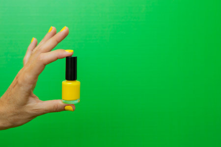 Horizontal photograph of a woman's hand with unpainted nails holding a bottle of yellow nail polish in front of a green background.の写真素材