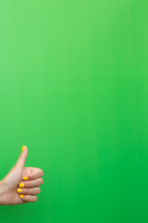 A woman's hand showing a thumbs up with her fingernails painted yellow on a green background.の写真素材
