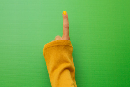 Hand of a woman with yellow painted fingernails on a green background raising her index finger in a mustard-colored shirt.の写真素材