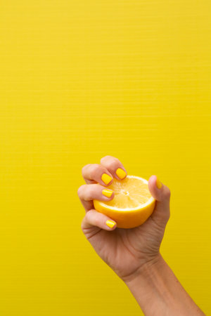 Hand of a woman with yellow fingernails holding half a lemon with yellow background