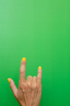 Hand of a woman with yellow painted nails on a green background doing the heavy metal horns.
