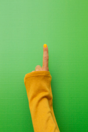 Hand of a woman with yellow painted fingernails on a green background raising her index finger in a mustard-colored shirt.の写真素材