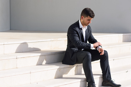Young businessman near a modern office building wearing black suit and white shirt sitting on the floor. Man with blue eyes in urban backgroundの写真素材