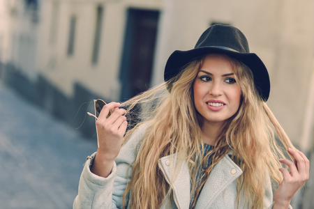 Portrait of beautiful young woman playing with her curly hair and smiling in urban background. Girl wearing jacket and hat.の写真素材