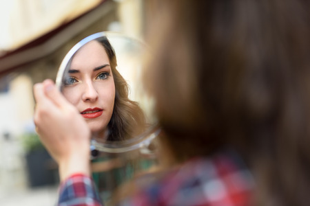 Portrait of young woman looking at herself in a little mirror in urban background.