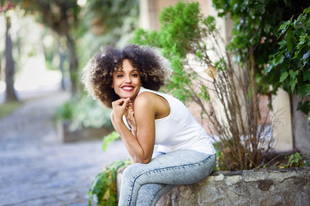 Young black woman with afro hairstyle smiling in urban park. Mixed girl wearing casual clothes sitting in the street.の写真素材