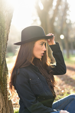 Portrait of thoughtful woman sitting alone outdoors wearing hat. Nice backlit with sunlightの写真素材