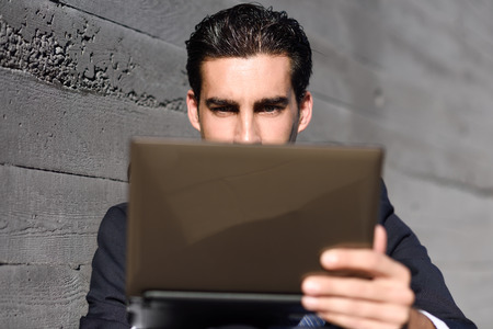 Good looking businessman using a laptop computer sitting in the street. Man wearing blue suit and tie in urban background.の写真素材