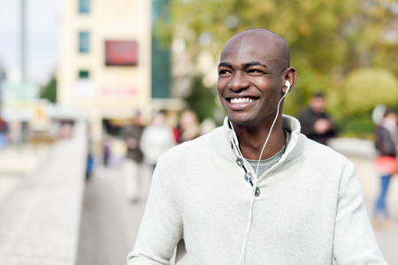 Black young man with a smartphone in his hand in urban background. Young african guy with shaved head wearing casual clothes and white headphones.の写真素材