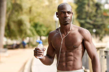 Fit shirtless young black man drinking water after running in urban background. Young male exercising with naked torso listening to music with headphones.の写真素材