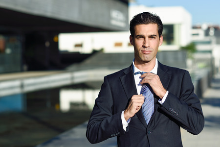 Young handsome businessman, dressed in blue suit, adjusting a tie in urban background with modern office buildings. Caucasian man with blue eyes.の写真素材