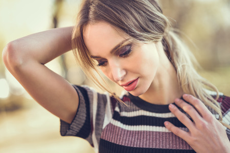 Close-up portrait of pensive young blonde woman standing in the street. Beautiful girl in urban background wearing striped sweater. Female with straight hair.の写真素材