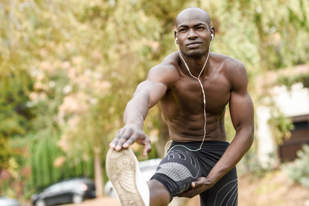 Fit shirtless young black man doing stretching before running in urban park. Young male exercising with naked torso listening to music with headphones.の写真素材