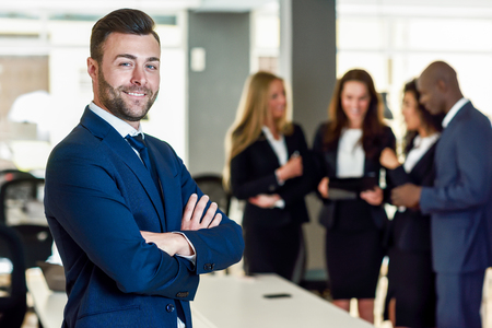 Caucasian businessman leader looking at camera in modern office with multi-ethnic businesspeople working at the background. Teamwork concept. Young man with beard wearing blue suit.の写真素材