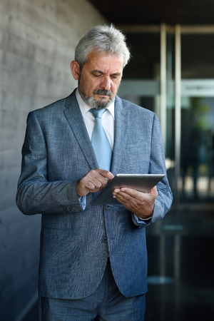 Portrait of a senior businessman with tablet computer outside of modern office building. Successful business man in urban background.の写真素材