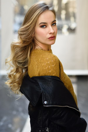 Blonde woman in urban background. Beautiful young girl wearing black leather jacket and mini skirt standing in the street. Pretty russian female with long wavy hair hairstyle and blue eyes.の写真素材