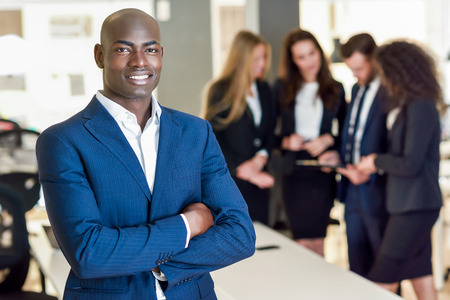 Black businessman leader looking at camera in modern office with multi-ethnic businesspeople working...