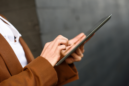 Young businesswoman with tablet computer standing outside of an office building. Beautiful woman wearing formal wear. Young girl with brown jacket.の写真素材