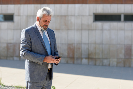 Portrait of a senior businessman texting with a smart phone outside of modern office building. Successful business man in urban background.の写真素材