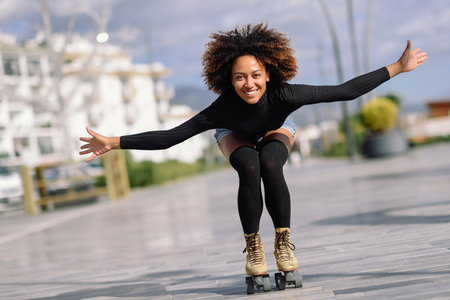 Young fit black woman on roller skates riding outdoors on urban street with open arms. Smiling girl with afro hairstyle rollerblading on sunny dayの写真素材