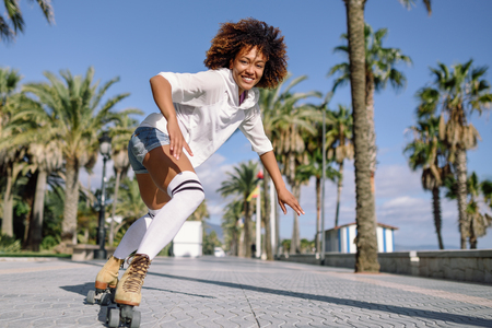 Smiling black woman on roller skates riding outdoors on beach promenade with palm trees. Smiling girl with afro hairstyle rollerblading on sunny day.の写真素材