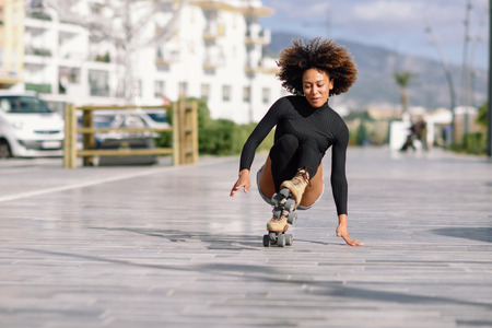 Young fit black woman on roller skates riding outdoors on urban street. Smiling girl with afro hairstyle falling to the ground.の写真素材