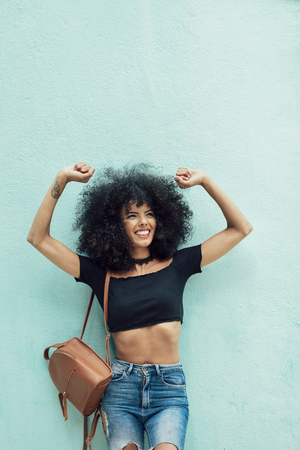 Smiling black woman with curly hair raising arms outdoors. Female wearing casual clothes in urban background. Lifestyle conceptの写真素材