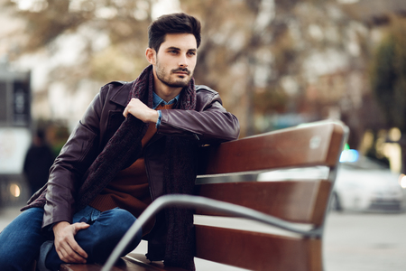 Thoughtful young man sitting on an urban bench. Attractive guy with modern hairstyle with lost look in the street.の写真素材