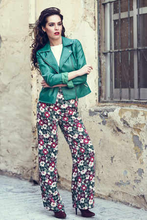 Young brunette woman, model of fashion, wearing green modern jacket and flower pants. Pretty caucasian girl with long wavy hairstyle. Female with red lips in urban background.の写真素材